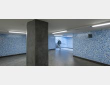 Unterirdischer Fußgängertunnel mit hellblauen Mosaikwänden und grauen Bodenfliesen. Eine Person geht eine Treppe hinauf, die aus dem Tunnel führt, während ein grauer Pfeiler im Vordergrund zu sehen ist.