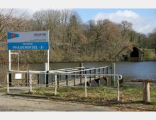 Anlegestelle mit einem Schild „Station Pfaueninsel 2“ und einem Steg, der ins Wasser führt, umgeben von Bäumen. Im Hintergrund ist ein hölzernes Bootshaus am Ufer sichtbar.
