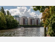 Moderne Wohngebäude mit großen Glasfassaden und braunen sowie beigen Fassadenelementen stehen entlang eines Flusses. Im Vordergrund ist ein kleines Segelboot auf dem Wasser zu sehen, umgeben von dichtem Grün am Flussufer.