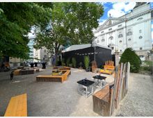 Ein Außenbereich mit Holzbänken und Tischen auf einem Kiesplatz, flankiert von Bäumen. Im Hintergrund steht ein weißes, historisches Gebäude mit verzierter Fassade, sowie eine schwarze Bühne mit Dekoration aus Topfpflanzen.