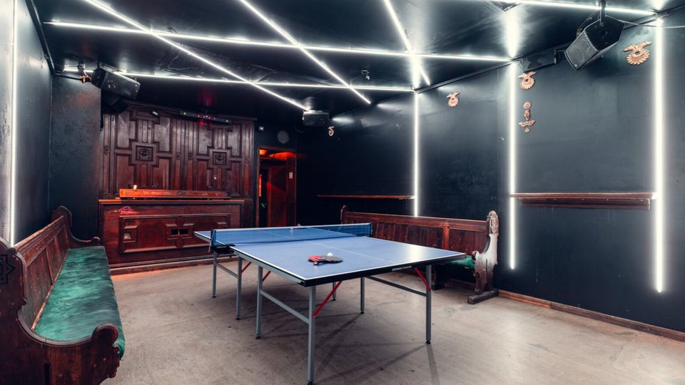 Innenraum eines dunklen Freizeitraums mit Tischtennisplatte in der Mitte, Holzbänken an den Seiten und einer Holztheke an der Rückwand. Decke und Wände sind mit hellen LED-Lichtstreifen in geometrischen Linienmustern und vertikalen Lichtleisten akzentuiert.
