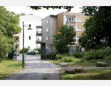 Ein Gehweg in einem kleinen Park, umgeben von Grünflächen, Sträuchern und Bäumen, führt auf eine Wohnstraße mit mehrstöckigen Gebäuden. Im Hintergrund sind ein helles und ein braun-beiges Wohnhaus mit Balkonen sowie parkende Autos sichtbar.