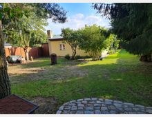 Garten mit Rasenfläche, Bäumen und Sträuchern, im Hintergrund ein gelbes Gartenhaus mit Flachdach und gemauertem Kamin. Ein gepflasterter Weg im Vordergrund und Holzzäune zur Eingrenzung des Grundstücks.