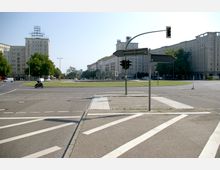 Straßenkreuzung mit Ampeln und Straßenmarkierungen in einer urbanen Umgebung. Im Hintergrund befinden sich große, symmetrische Wohngebäude im Stil des sozialistischen Klassizismus, umgeben von Bäumen und Grünflächen.