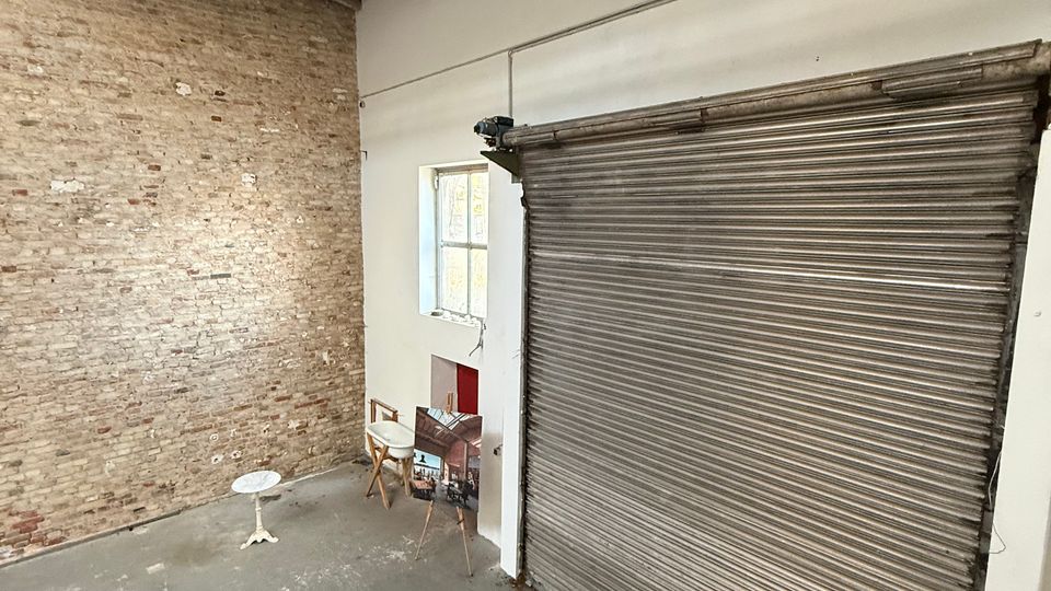 Innenraum einer hohen Lager- oder Werkstatthalle mit sichtbaren Deckenbalken, Backsteinwand und grauem Betonboden. Rechts befindet sich ein großes metallisches Rolltor neben einem kleinen Fenster in einer weißen Wand.