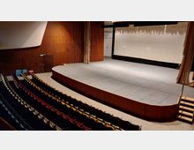 Innenansicht eines Theatersaales mit einer erhöhten Bühne, umgeben von hölzernen Wandverkleidungen. Vor der Bühne befinden sich gestaffelte Sitzreihen mit verschiedenfarbigen Polstern, während auf der Bühne ein heller Vorhang zur Seite gezogen ist.