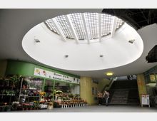 Ein unterirdischer Durchgang mit einer ovalen Lichtkuppel aus Glasgittern, darunter ein Blumenladen mit Pflanzen und bunten Blumenarrangements. Rechts sind Treppen mit gelben Fliesen an der Wand sichtbar, die nach oben ins Freie führen.