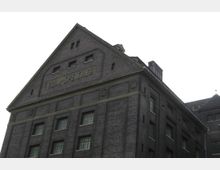 Backstein-Gebäude mit der Aufschrift „Zoll-Speicher“ auf dem Giebel, rechteckige Fenster mit Gitterelementen und steilem Dach; Architektur im industriellen Stil vor bewölktem Himmel.