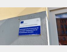 Eine Infotafel an einer Gebäudewand mit der Aufschrift: „Denkmalgerechte Erneuerung der ehemaligen Fabrikantenvilla der Rohtabakfabrik Vierraden“. Im oberen Bereich befinden sich das EU-Logo und das Logo des Landes Brandenburg. Rechts daneben ein Fenster mit Spiegelung eines Ziegelgebäudes.