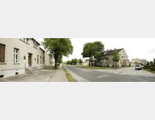 Straßenszene in einer Wohngegend mit großen Bäumen entlang des Gehwegs, einfachen, zweigeschossigen Häusern aus Putz und vereinzelt stehenden Autos. Die Straße ist asphaltiert und führt durch eine ruhige Umgebung.