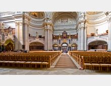 Innenansicht einer prunkvollen Kirche mit hohen, verzierten Säulen, reichlich dekorierten Gewölbedecken und hölzernen Kirchenbänken, die symmetrisch angeordnet sind. Im Hintergrund befindet sich eine kunstvolle Empore mit vergoldeten Elementen und drei großen Doppeltüren.