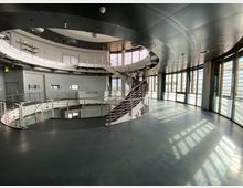 Innenansicht eines modernen Gebäudes mit einer runden Galerieebene, einer geschwungenen Treppe aus Metallgeländern und großen Fenstern, die viel Tageslicht hereinlassen. Die Decke ist dunkel und verläuft bogenförmig, während der Boden aus glattem dunklem Material besteht.