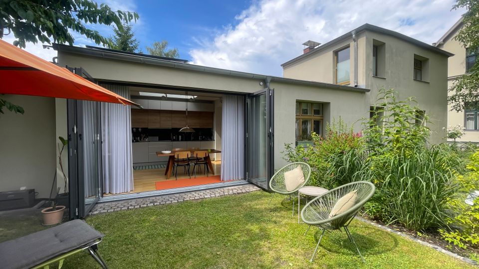 Moderner Wohnhaus-Anbau mit großer, aufgeklappter Glasfront zur Terrasse, die den Blick in eine offene Küche mit Esstisch freigibt. Im Vordergrund liegt ein Rasen mit zwei Lounge-Stühlen und einem Sonnenschirm, daneben ein dicht bepflanztes Gartenbeet.