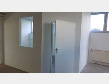 Innenraum mit weißen Wänden, einer halb geöffneten Tür, einem kleinen Fenster mit Glasscheibe und einem Heizkörper an der Wand unter einem weiteren rechteckigen Fenster. Der Boden ist unbehandelt.