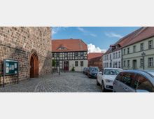 Gepflasterter Platz mit historischen Gebäuden, darunter eine Fachwerkfassade und eine steinerne Kirchenwand mit einem gotischen Torbogen. Am Rand des Platzes parken Autos, und es sind Laternen sowie ein Schaukasten sichtbar.