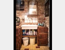 Rustikale Waschküche mit freiliegender Backsteinwand, einem weißen Keramikwaschbecken und einem goldgerahmten Wandspiegel darüber. Darunter befinden sich Regale mit Putzmitteln sowie zwei Eimer, flankiert von hölzernen, gealterten Schränken.