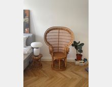 Ein heller Raum mit einem markanten, geflochtenen Rattansessel im Vordergrund. Daneben stehen ein kleiner Beistelltisch mit einer weißen Papierlampe sowie eine grüne Zimmerpflanze auf einem Stapel Bücher. Der Boden besteht aus hellem Fischgrätparkett.