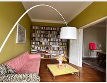 Wohnzimmer mit grün gestrichenen Wänden und Holzboden. Im Raum stehen ein gemusterter roter Sofa, ein gelber Couchtisch mit Dekoration und eine große weiße Bogenlampe. Im Hintergrund befindet sich eine große Bücherwand, und eine offene Tür führt zu einem angrenzenden Raum.