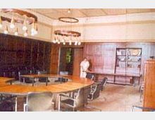 Ein Tagungsraum mit einem großen, ovalen Holztisch im Zentrum, umgeben von schwarzen Stühlen. Die Wände sind mit Holzvertäfelungen verkleidet, und an der Decke hängen runde Kronleuchter mit mehreren Glühbirnen.