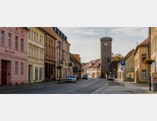 Straßenszene in einer kleinen Stadt mit historischen Gebäuden, darunter ein Uhrturm aus Naturstein im Hintergrund. Die Straße ist gesäumt von bunt verputzten Häusern mit dekorativen Fassaden, geparkten Autos und Laternen.