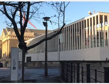 Moderne Fassade des Deutschen Historischen Museums in Berlin mit weißen Säulen und einem weitläufigen Glasdach, daneben ein klassizistisches Gebäude mit Säulen und Zierelementen. Im Vordergrund stehen ein Baum und ein Wegweiser.
