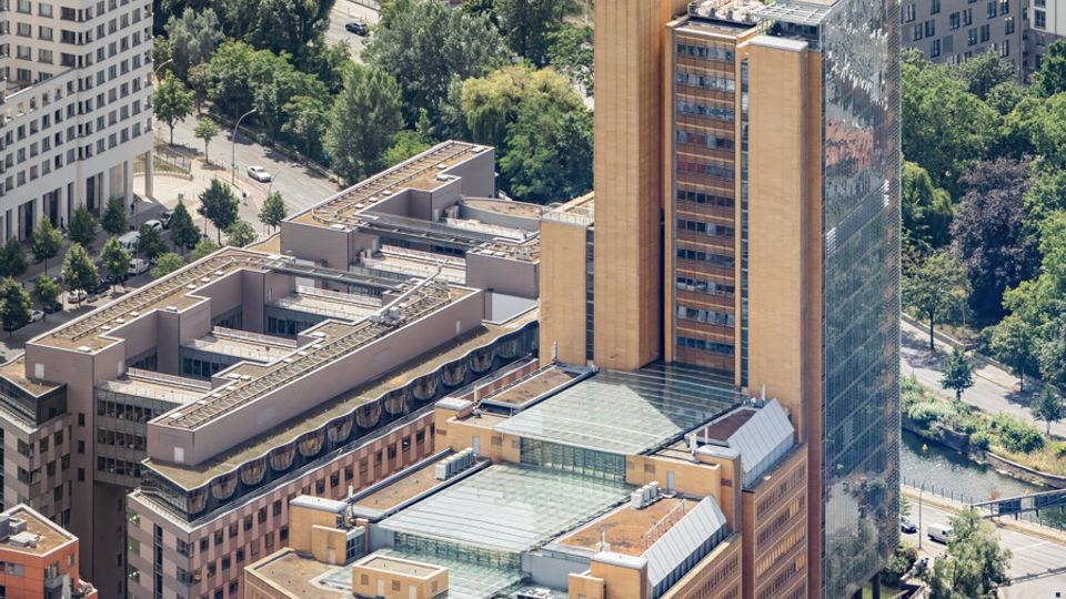 Aerial view of a large urban office complex with tan modern buildings, a tall narrow tower topped with green glass, and a taller dark glass high-rise beside it. Surrounding streets, trees, and adjacent buildings—including rail tracks and a green park area—frame the site.