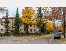 Wohnstraße in herbstlicher Umgebung mit mehrstöckigem Wohnhaus, hellen Fassaden und rotem Ziegeldach. Die Straße ist gesäumt von Bäumen mit bunt gefärbtem Herbstlaub, einem Gehweg, parkenden Autos und einem Holzzaun entlang der Grundstücke.
