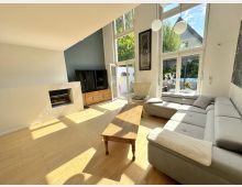 Heller, moderner Wohnbereich mit hohen Fensterfronten und Terrassentür, die viel Tageslicht auf den hellen Holzboden lässt. Zu sehen sind ein L-förmiges Sofa, ein Holztisch, ein Kamin, ein Fernseher auf Sideboard sowie Blick in den begrünten Außenbereich.