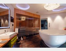 Modernes Badezimmer mit großer, freistehender Badewanne, einem Waschbecken auf einer Holzablage mit grünen Schubladen und einer verglasten Sauna im Hintergrund. Der Raum hat helle Wände, eine hängende Designer-Leuchte und braunen Fliesenboden.