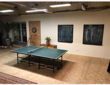 Ein Indoor-Freizeitraum mit einer Tischtennisplatte in der Mitte, einer Dartscheibe an einer Holzplatte an der Wand und zwei Tafeln mit notierten Ergebnissen. Im Hintergrund befinden sich eine Topfpflanze, ein kleines Möbelstück und eine Tür mit Glaseinsätzen.
