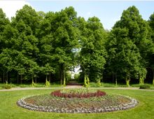Ein gepflegter Park mit einer kreisförmigen Blumenanlage im Vordergrund, umgeben von grünem Rasen. Im Hintergrund stehen hohe, dicht belaubte Bäume, durchzogen von einem Weg, der zu einem hellen Gebäude in der Ferne führt.