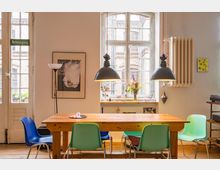 Heller Innenraum eines Wohnbereichs mit einem Holztisch und bunten Stühlen, großen Fenstern mit Sprossen, einem Heizkörper an der Wand und dekorativen Elementen wie einer Lampe, Pflanzen und Bildern. Im Hintergrund ist durch die Fenster eine Backsteinfassade sichtbar.