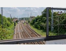 Blick auf eine Eisenbahnstrecke mit mehreren Gleisen, umgeben von Grünflächen und vereinzelten Gebäuden im Hintergrund. Im Vordergrund ist ein Metallgitter eines Brückengeländers zu sehen, während im Hintergrund ein kleiner Bahnhof und Oberleitungen sichtbar sind.