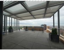 Rooftop terrace with large square floor tiles, a metal slatted canopy overhead, and glass-and-metal railings along the edges. Planters with shrubs line the perimeter, with a wide city skyline view beyond.