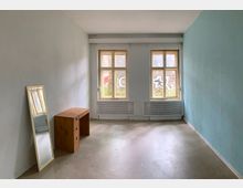 Unmöbliertes Zimmer mit zwei großen Fenstern, die durchscheinendes Tageslicht hereinlassen; an der linken Wand steht ein bodenstehender Spiegel und ein kleiner Holzschreibtisch. Die Wände sind weiß und hellblau gestrichen, der Boden ist aus Beton, und draußen ist eine Backsteinmauer mit Graffiti sichtbar.