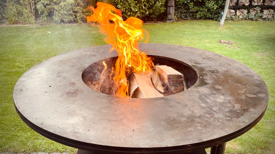 Runder, metallener Feuerkorb mit breiter Grillplatte und zentraler Öffnung, in der Holzscheite mit sichtbaren Flammen brennen. Dahinter ein Garten mit Rasenfläche, dichter Heckenbepflanzung und einem Holzlagerregal am Rand.