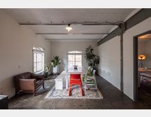 Heller Raum mit minimalistischer Einrichtung, bestehend aus einem weißen Tisch, farbigen Stühlen, einer Holzbank, einem Ledersessel und großen Topfpflanzen. Sichtbare Stahlträger und ein weiß gestrichener Backsteinhimmel sowie Fenster mit Blick nach draußen betonen das industrielle Design.