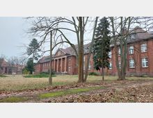 Rotes, historisches Backsteingebäude mit Säulenportal und Satteldach, umgeben von einer Wiese mit kahlen Bäumen im Vordergrund. Im Hintergrund sind weitere Backsteingebäude und einzelne Büsche sichtbar.