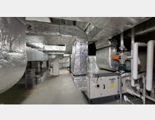 Technikraum mit komplexem Lüftungssystem, bestehend aus großen, silbern isolierten Rohren und technischem Equipment. Im Vordergrund befinden sich weitere Leitungen und Steuerungseinheiten, die den Raum dominieren.