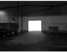 Schwarz-Weiß-Fotografie eines Industriegebäudes mit einer geöffneten Garagentür, durch die helles Licht scheint. Links im Bild steht ein geparktes Auto, während kahle Bäume und Rohrleitungen die Wandstruktur ergänzen.
