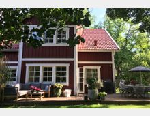 Zweistöckiges rotes Holzhaus mit weißen Fensterrahmen und einem roten Ziegeldach, umgeben von Bäumen. Im Vordergrund eine Holzterrasse mit Sitzmöbeln, Pflanzen in Töpfen und einem Esstisch mit Sonnenschirm.