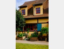 Fachwerkhaus mit gelber Fassade, roten Fenstern und blauen Klappläden, umgeben von Hochbeeten mit Pflanzen und Blumen. Der Vorgarten ist bepflanzt, mit Gehweg und angrenzendem Rasen.