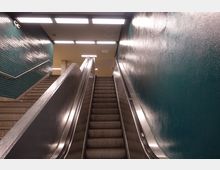 Rolltreppe in einer U-Bahn-Station mit grün gekachelter Wand auf der linken Seite und glatt gestrichener Wand auf der rechten Seite. Über der Rolltreppe befinden sich rechteckige Deckenlampen, die den Bereich beleuchten.