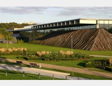Moderne Gebäudestruktur mit großer Glasfront und einer Wand aus diagonal angeordneten Holzstämmen, umgeben von einer gepflegten Parkanlage mit Rasenflächen, Blumenbeeten und Sitzbänken. Im Hintergrund sind Bäume und Teile einer hügeligen Landschaft erkennbar.
