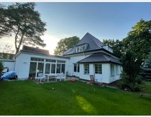 Ein modernes zweistöckiges Wohnhaus mit weißer Fassade und dunklem Satteldach, umgeben von einem gepflegten Garten mit Rasenfläche, Sitzmöbeln und vereinzelten Bäumen. Große Fensterfronten und eine Terrasse prägen die Rückseite des Gebäudes.