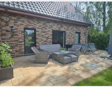 Gepflasterte Terrasse an einem Backsteinhaus mit Satteldach, Dachrinne und zwei Außentüren. Auf der Terrasse stehen eine Rattan-Sitzgruppe mit Tisch sowie mehrere Liegestühle; dahinter Gartenhecken und Bäume.