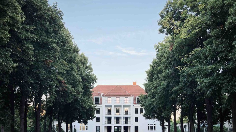 Weiße Villa mit rotem Ziegeldach, symmetrischer Fassade und Balkonen, umgeben von hohen Bäumen, die einen Weg durch einen grasbedeckten Garten einrahmen. Der Himmel ist klar und blau.
