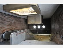 U-Bahn-Zugang mit roten Backsteinwänden, einer Rolltreppe und einem Treppenabgang mit Metallhandläufen; eine Oberlichte aus Holzlamellen und vier runde Fenster sorgen für Licht.