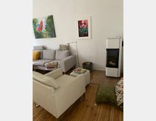 Wohnzimmer mit Holzboden, grauem Sofa, cremefarbenem Sessel und einem weißen Kaminofen. An der Wand hängen zwei gerahmte Bilder, eine grüne Sitzkissenmatte liegt auf dem Boden. Ein Beistelltisch mit Magazinen steht neben dem Sofa.