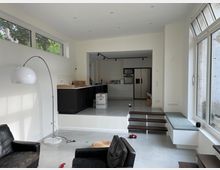 Helles modernes Wohnzimmer mit offenem Übergang zur Küche, erhöht durch eine kleine Treppe. Große Fenster lassen viel Licht herein, links steht eine weiße Bogenlampe, und die Küche zeigt schwarze Schränke und eine helle Arbeitsfläche.